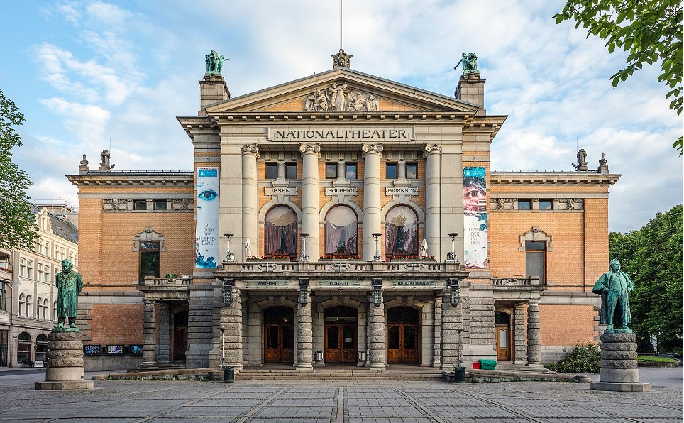 A large bulding in a light beige colour. Has the title 'NATIONALTHEATER' engraved into the building. There is a sculpture of two men standing to the right and left of the building.