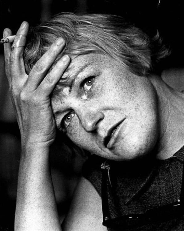 Black and white image of young woman with short, light-coloured hair holding her hand to her head with a cigarette in it.