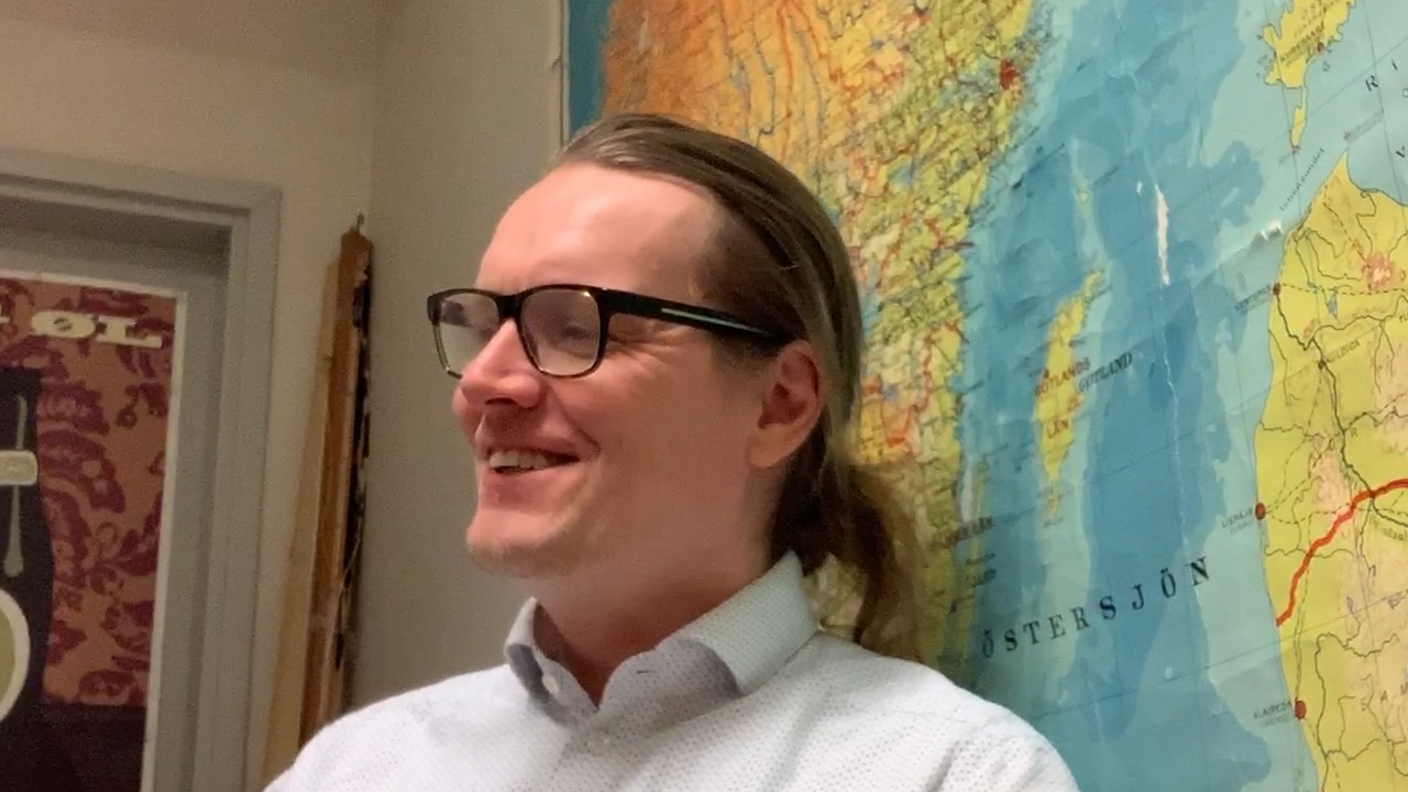 A smiling young man with long hair in a ponytail wearing glasses sitting in front of a world map